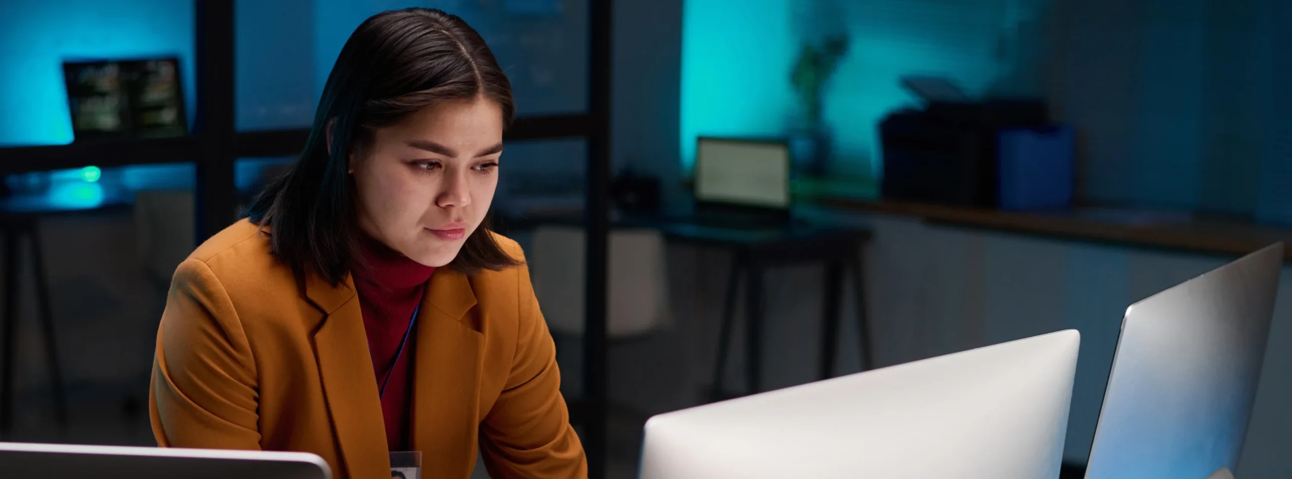 Woman in a mustard blazer working at a computer in a dimly lit office.