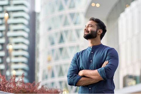 A confident man in a blue shirt stands outdoors, arms crossed, looking up in a modern urban setting.
