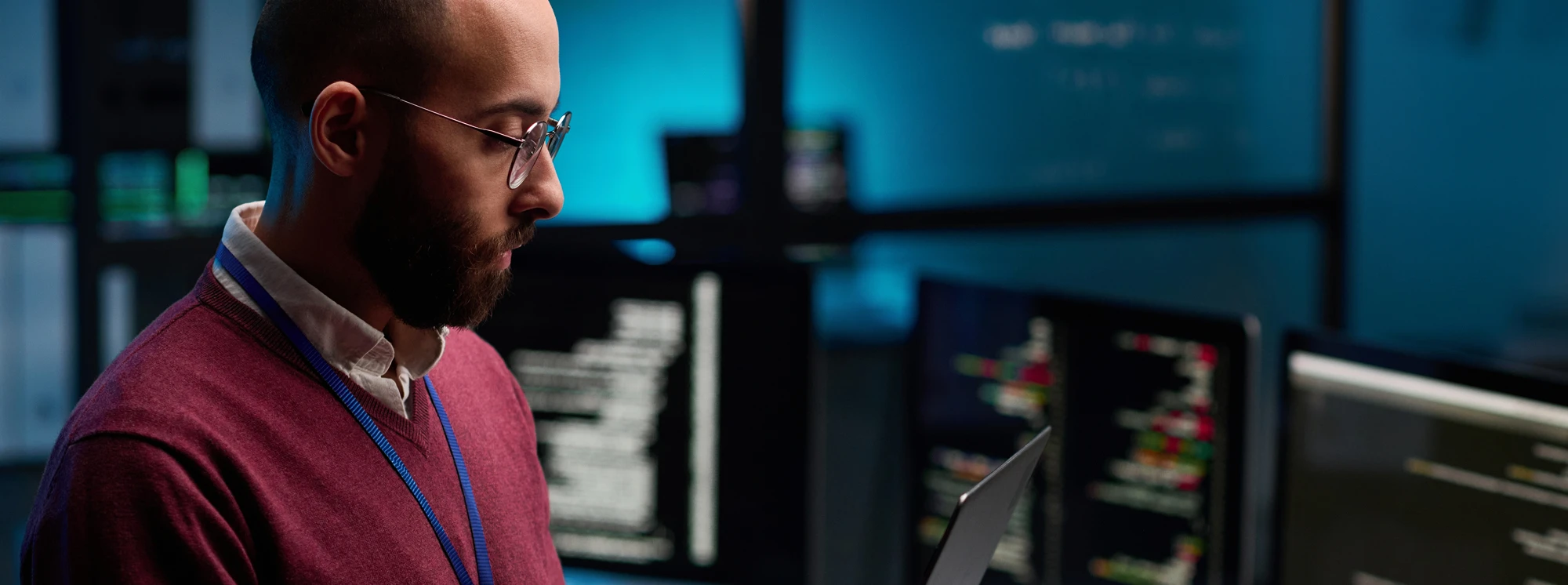 Man in glasses analyzing code on laptop, surrounded by monitors in a tech workspace.